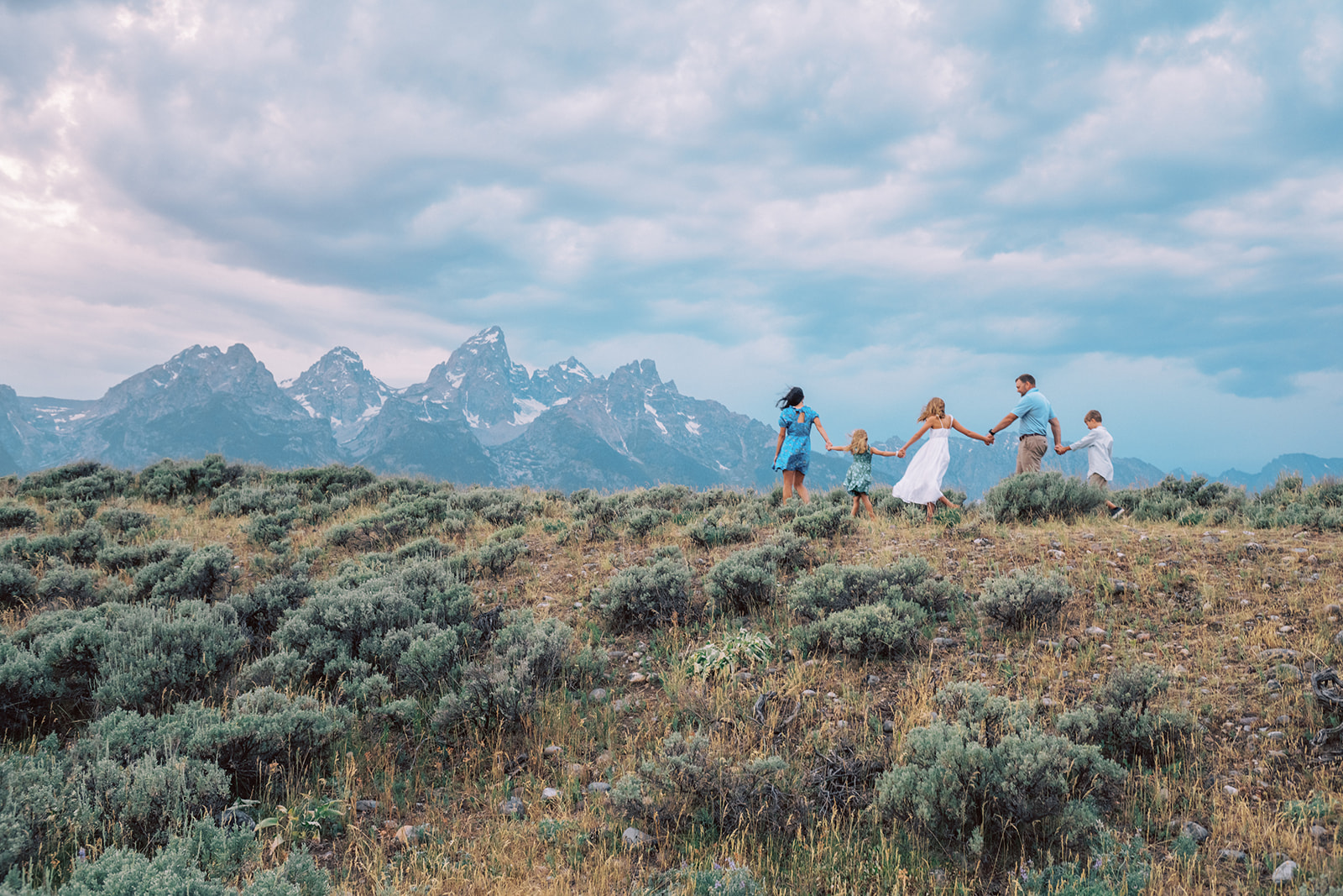 A mom and dad walk holding hands with their three young children on a ridge in white and blue after finding things to do in spring in Grand Teton National Park with kids