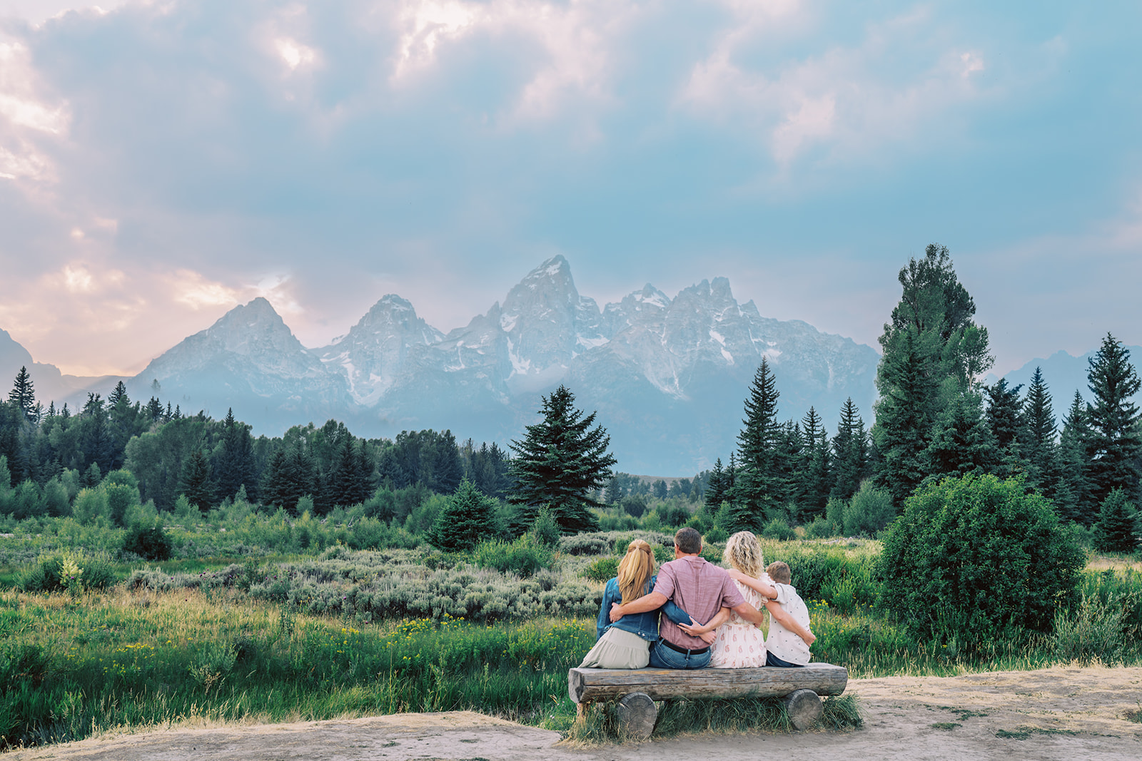 A happy family sit with arms locked on a wooden log bench by a pond admiring the majestic mountains