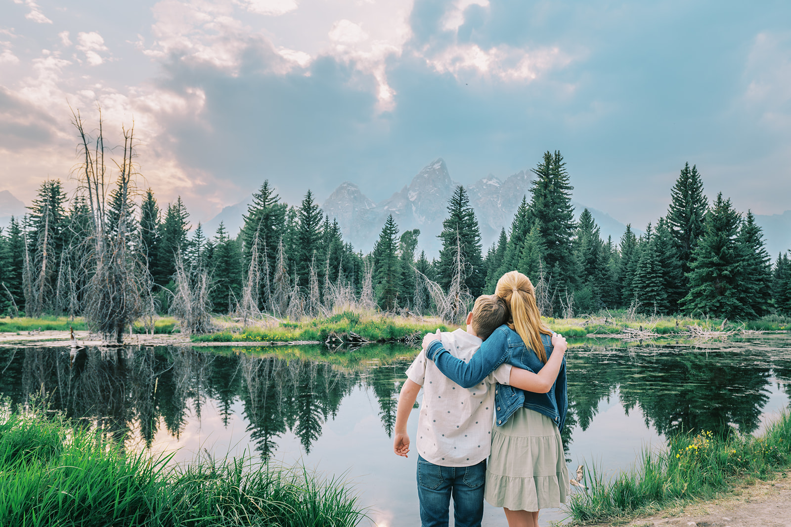 Teen brother and sister with arms around each other admire the mountains across a pond and behind a pine forest after finding things to do in summer in Jackson Hole
