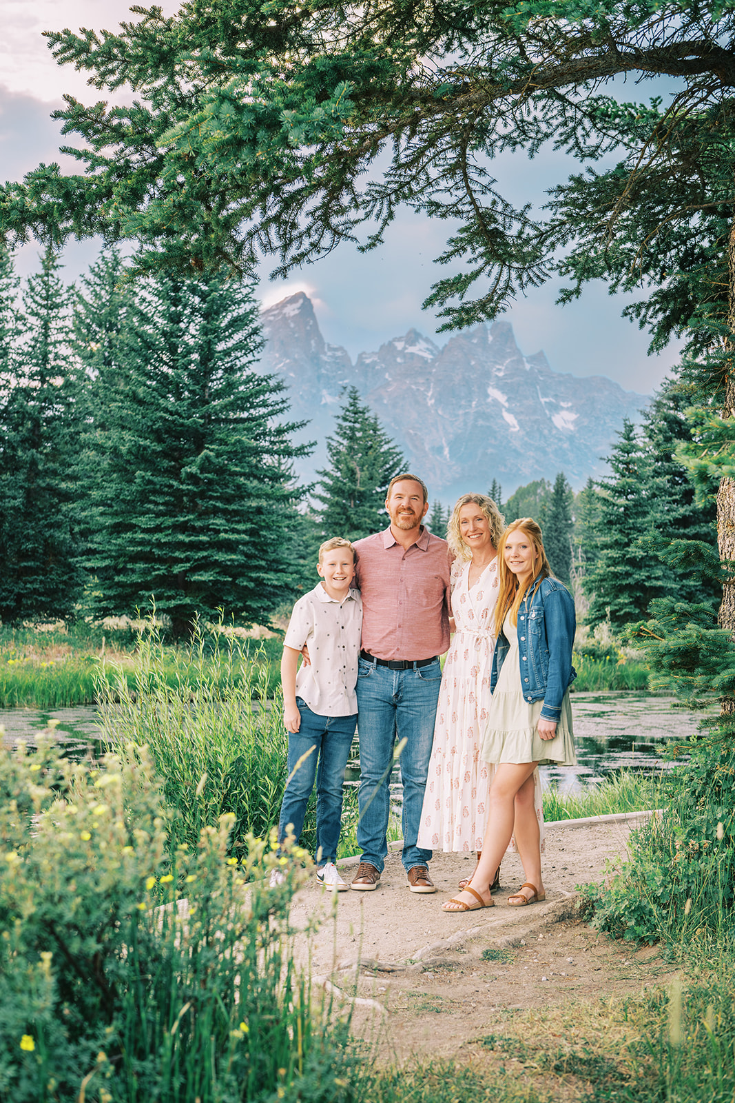 A happy mom and dad stand with their young son and teen daughter in a mountain trail smiling in front of a pond
