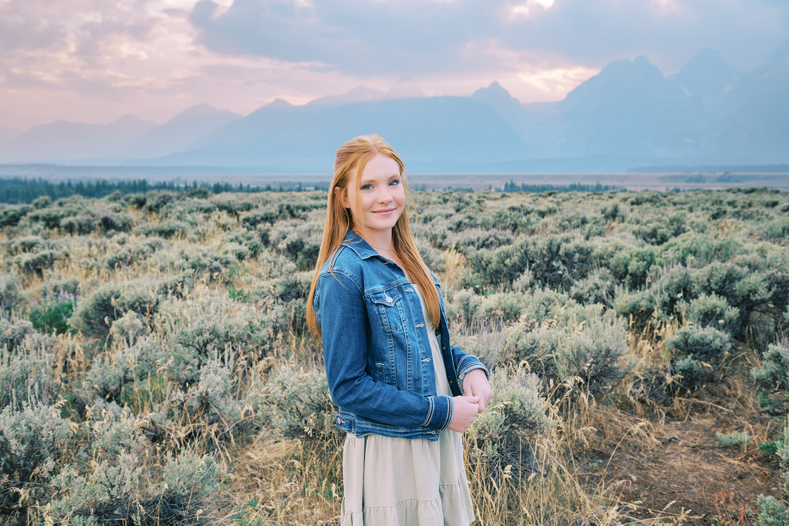 A smiling teenager in a green dress and denim jacket stands in a trail at sunset in a meadow after finding things to do in summer in Jackson Hole