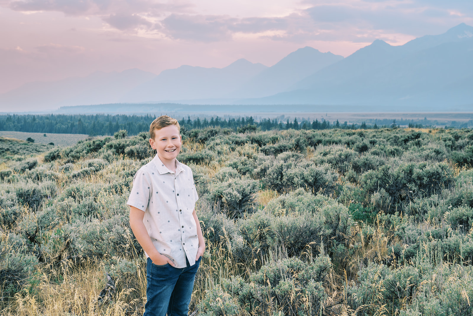 A young boy in a white button down and jeans stands smiling with hands in pockets in a mountain meadow during one of the things to do in summer in Jackson Hole