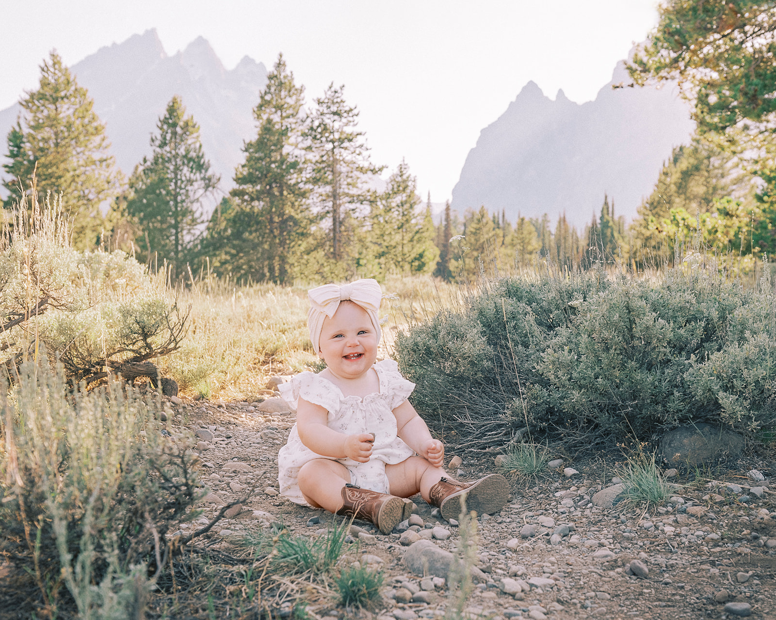 A giggling baby girl in a white dress and big bow smiles while sitting in a forest trail at sunset