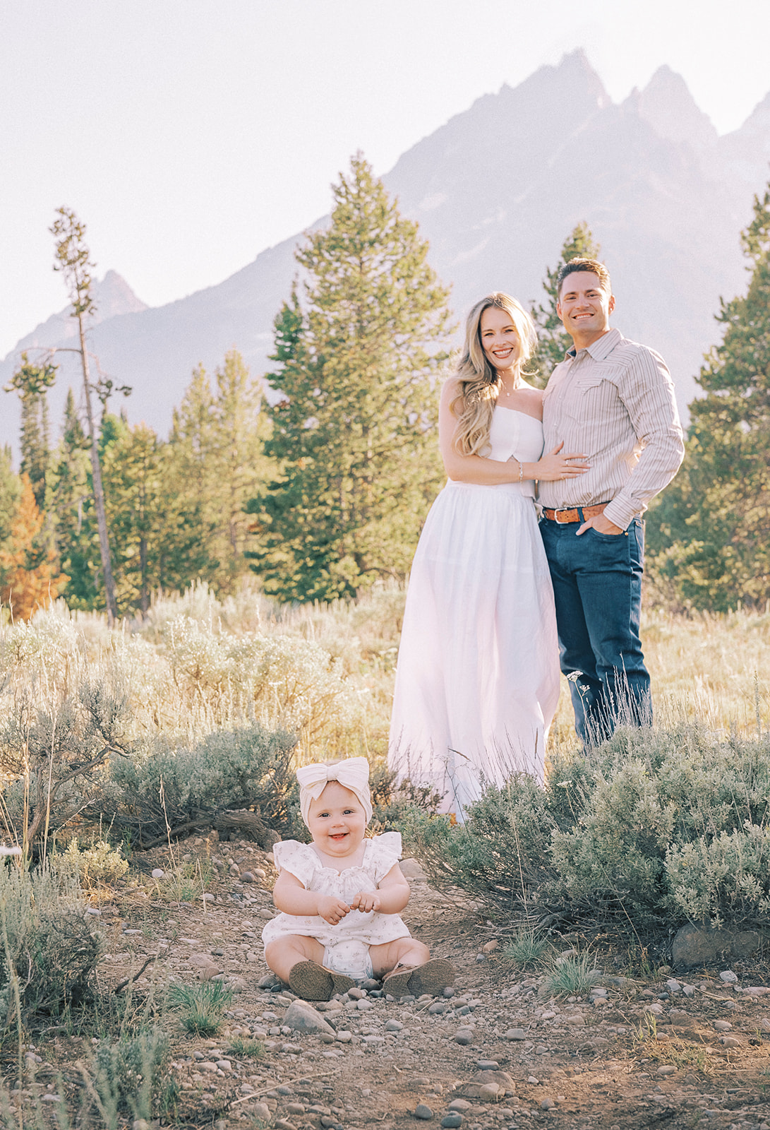 A toddler girl sits playing with rocks in a trail as mom and dad smile behind her after visiting Montessori School Of The Tetons