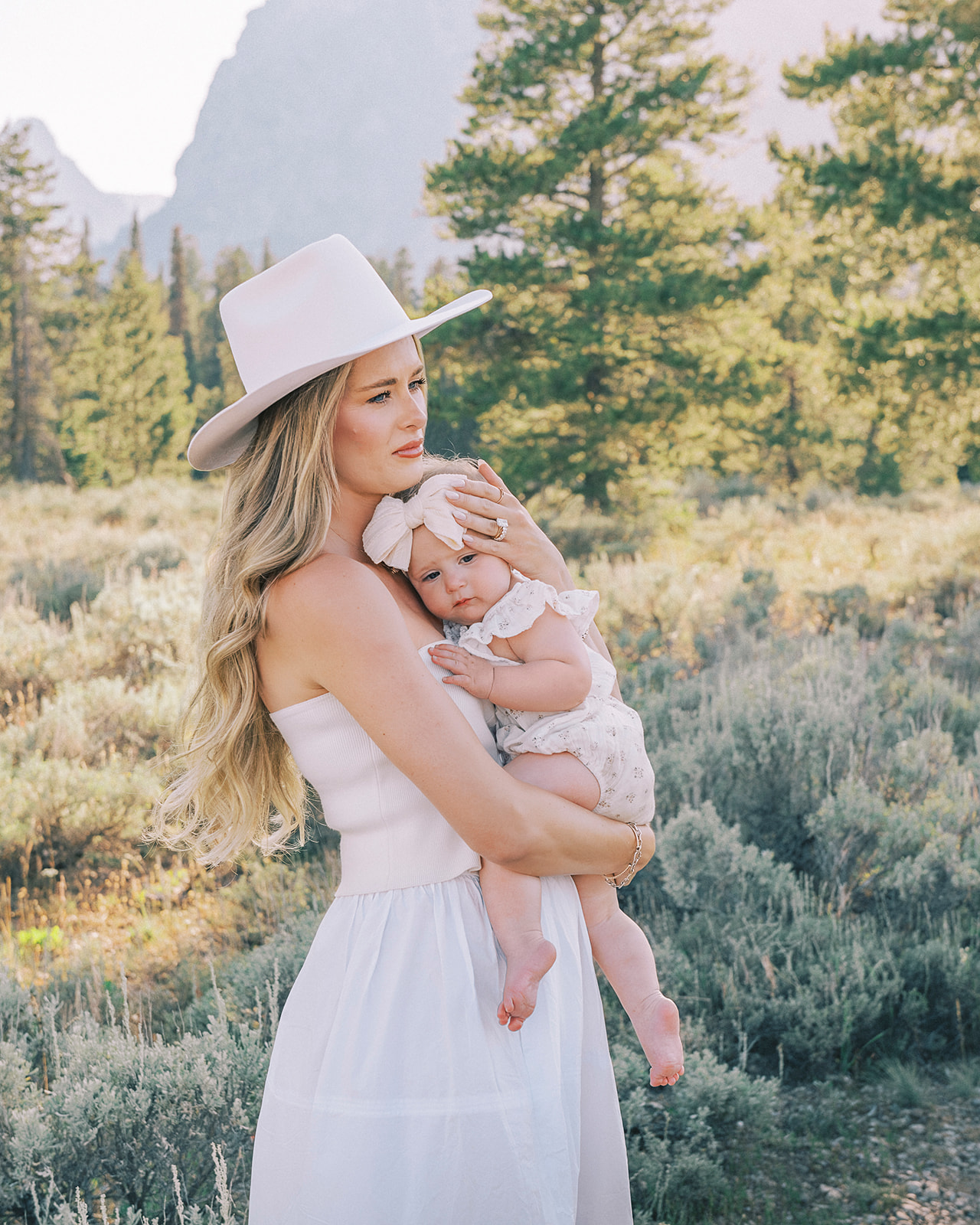 A mother in a white dress and cowboy hat cradles her baby girl against her chest in a mountain trail after visiting Montessori School Of The Tetons