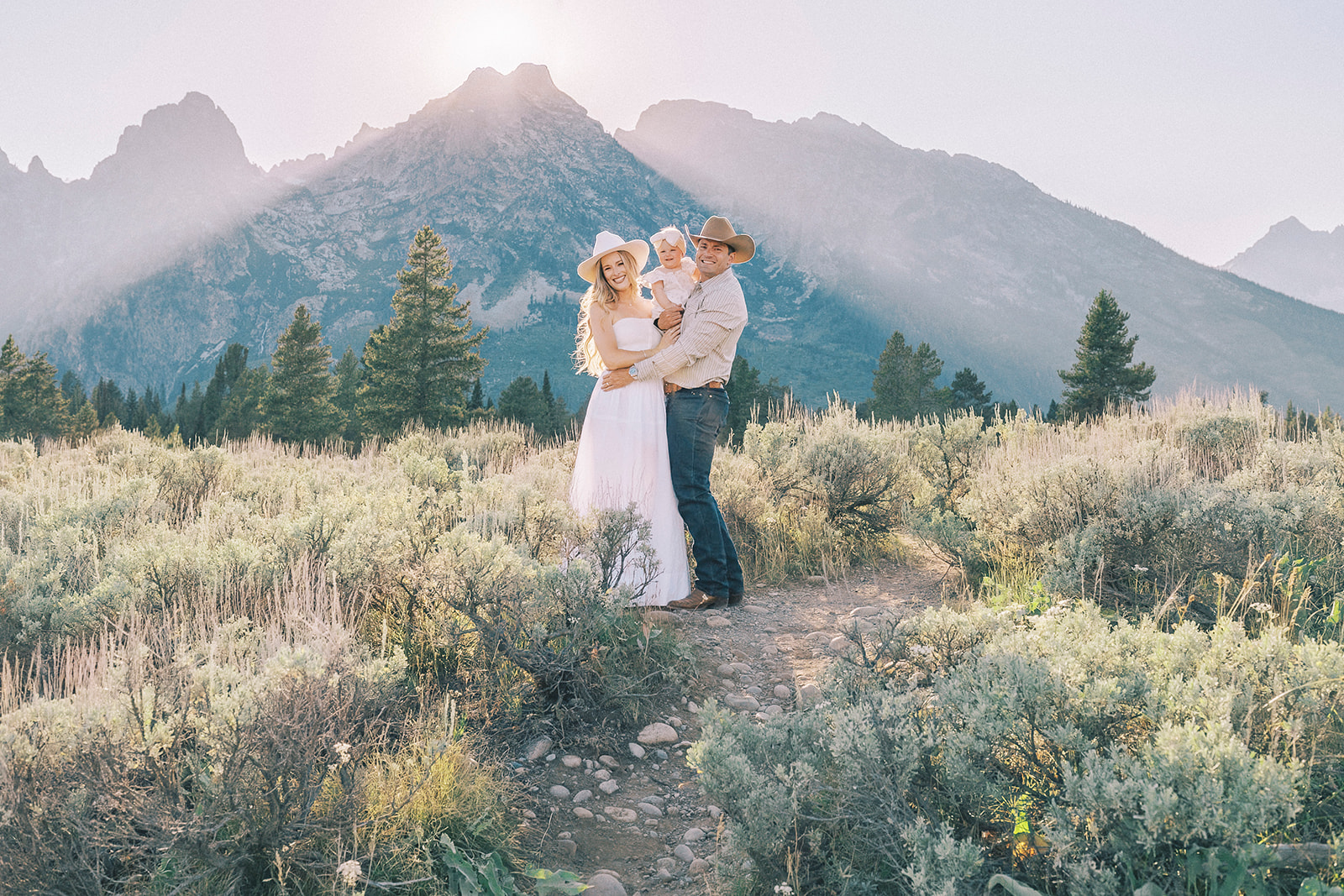 Mom and dad stand smiling in a meadow in cowboy hats holding their toddler daughter between them on a trail after exploring Montessori School Of The Tetons