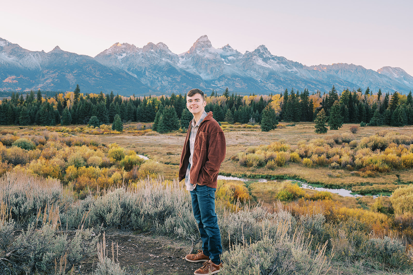 A smiling high school senior in a brown jacket and jeans stands in a trail at sunset after finding great tuxedo rental in Jackson, WY