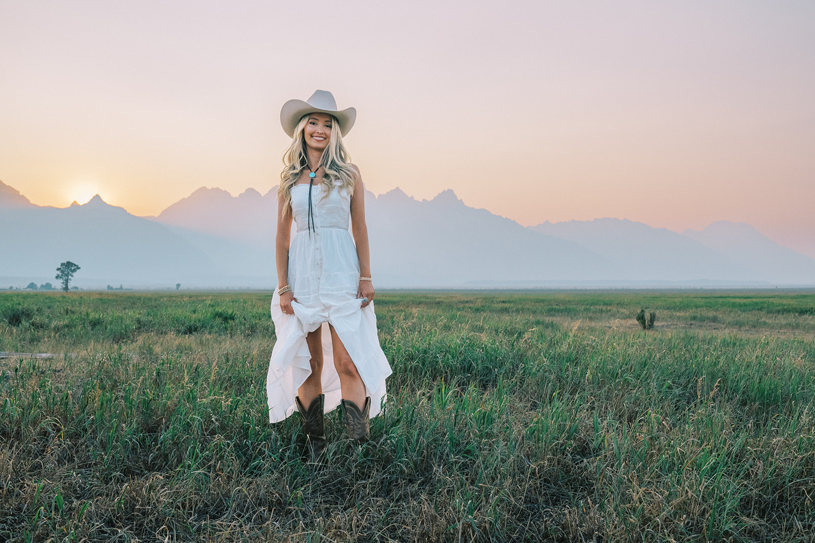 A happy graduate walks in a pasture at sunset in the tetons in a white dress and cowboy hat and boots