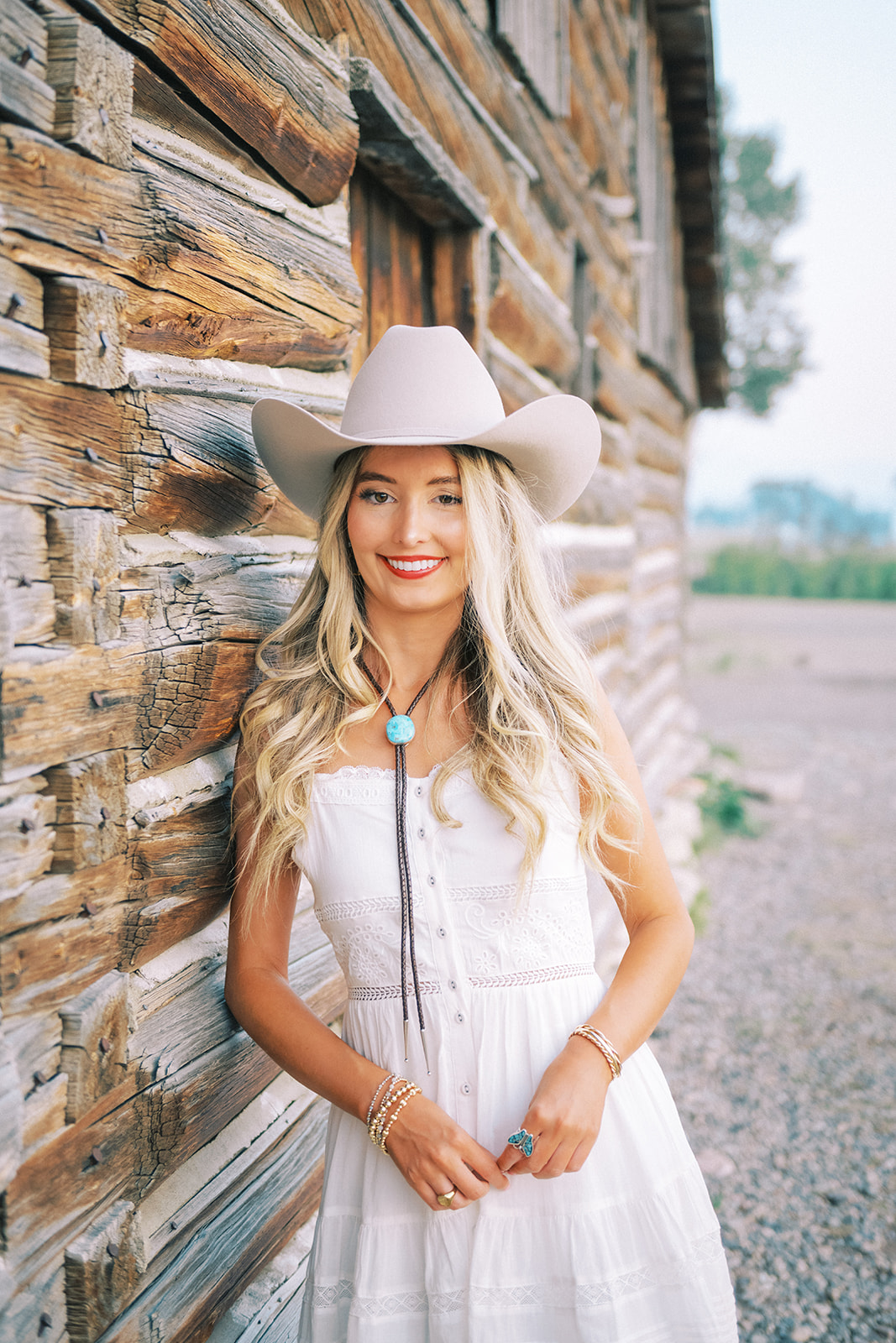 A happy high school senior in a white dress and cowboy hat leans against a log cabin at sunset smiling after finding tanning in Jackson, WY