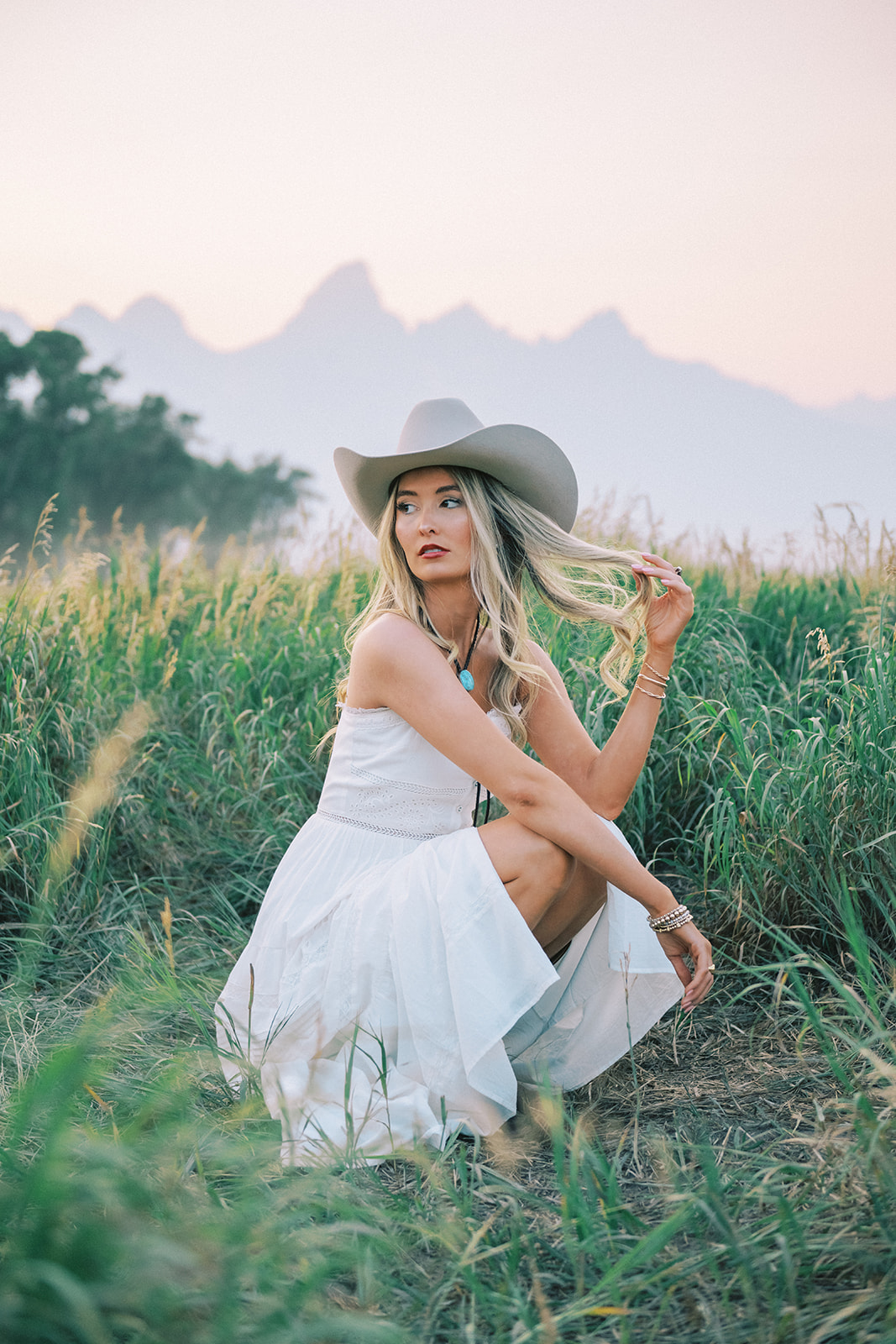 A high school senior in a white dress and tan cowboy hat twirls her hair while squatting in tall grasses at sunset after enjoying tanning in Jackson, WY